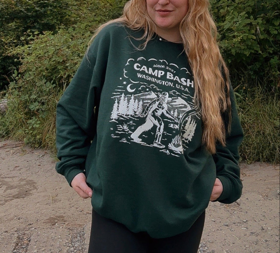 Person wearing a green sweatshirt with 'Camp Bash' design outdoors and bigfoot